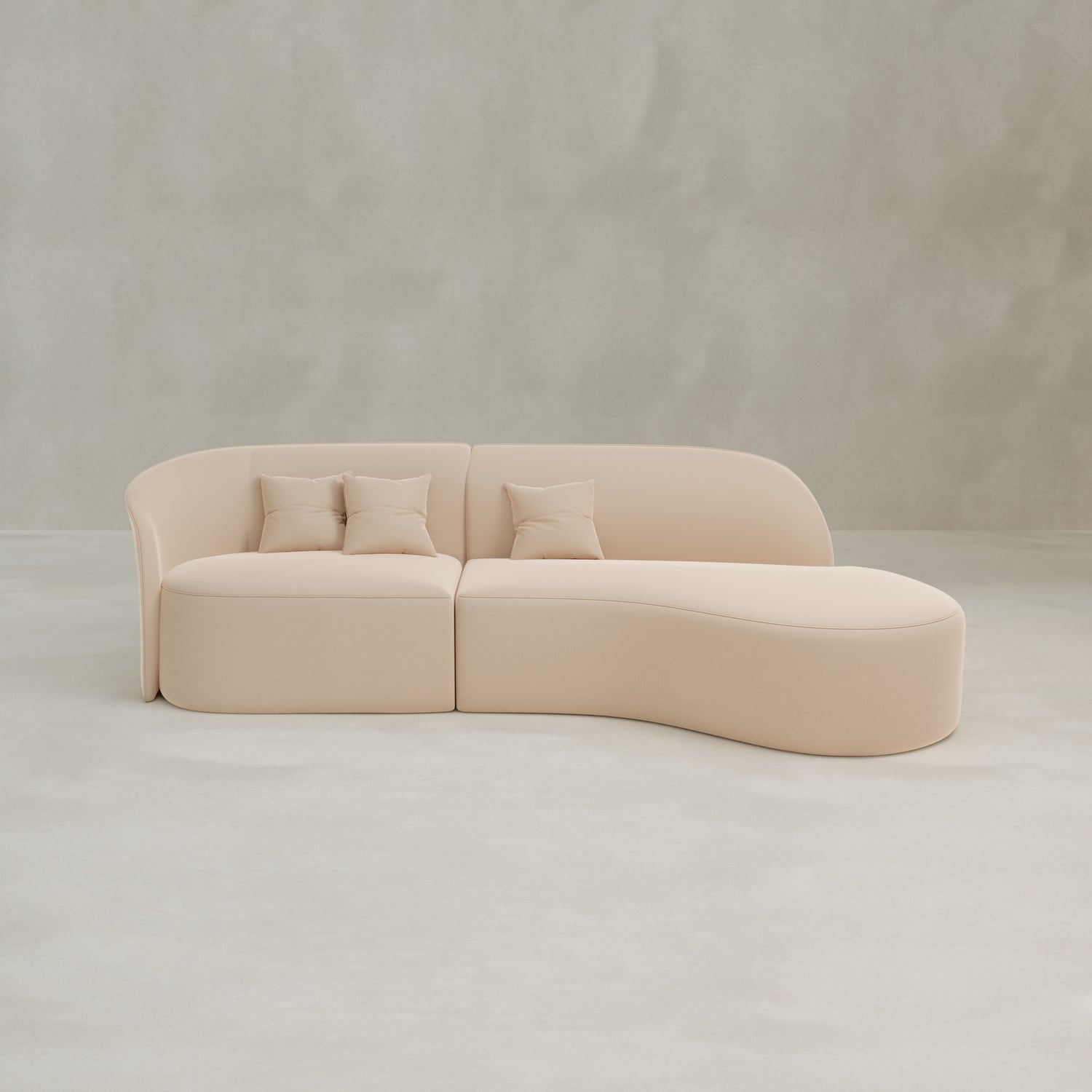 The Marlow Curved Modular Three Seater Sofa, featuring luxurious beige upholstery and three matching throw pillows, sits on a light gray floor against a plain, light-colored wall.
