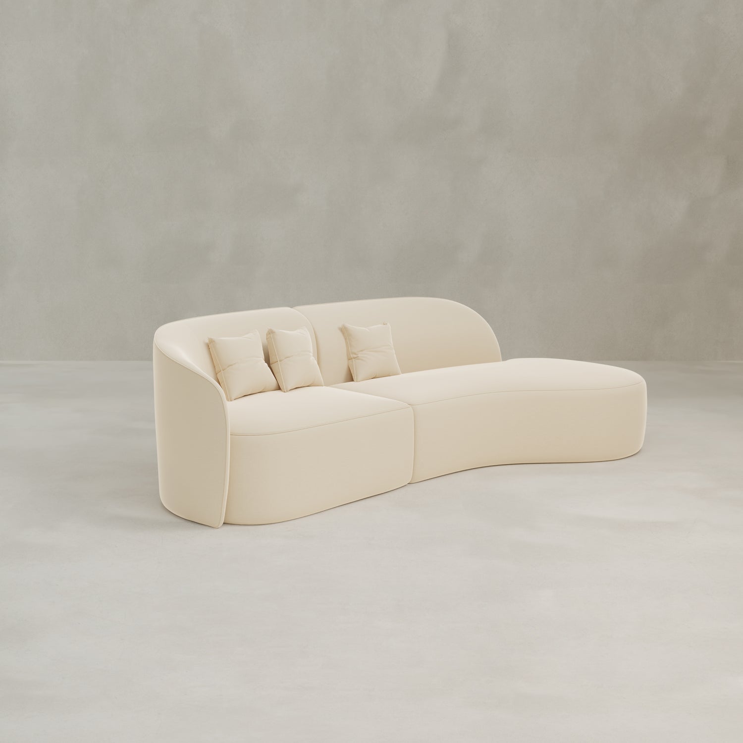 The Marlow Curved Modular Three Seater Sofa features modern cream-colored upholstery and comes with three matching throw pillows, shown in an empty room with light gray walls and a smooth, pale floor.
