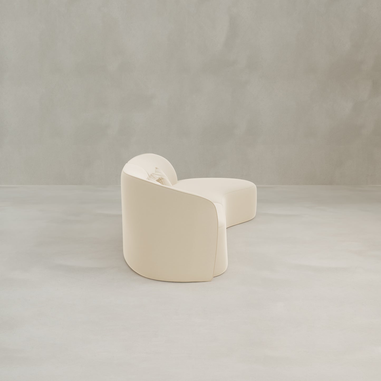 The Marlow Curved Modular Three Seater Sofa, in cream velvet with smooth upholstery, stands on a light gray concrete floor against a textured, light gray wall for a modern look.