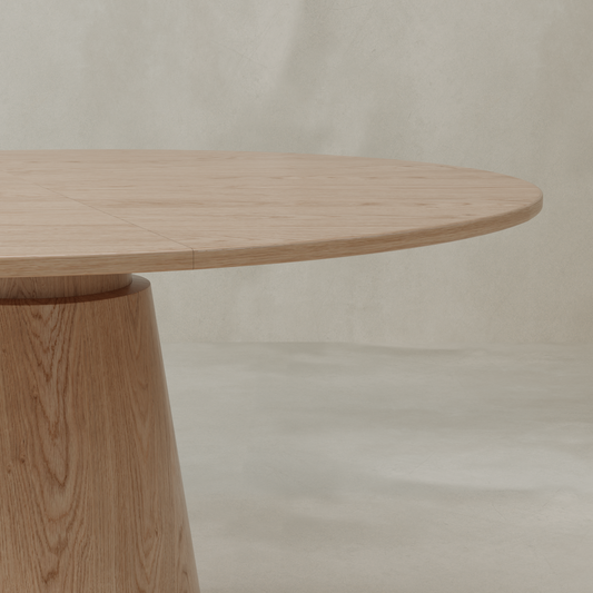 A close-up view of the Harlow Dining Table, featuring a smooth, natural wood finish and a thick, cylindrical pedestal base, set against a plain light beige wall and floor.