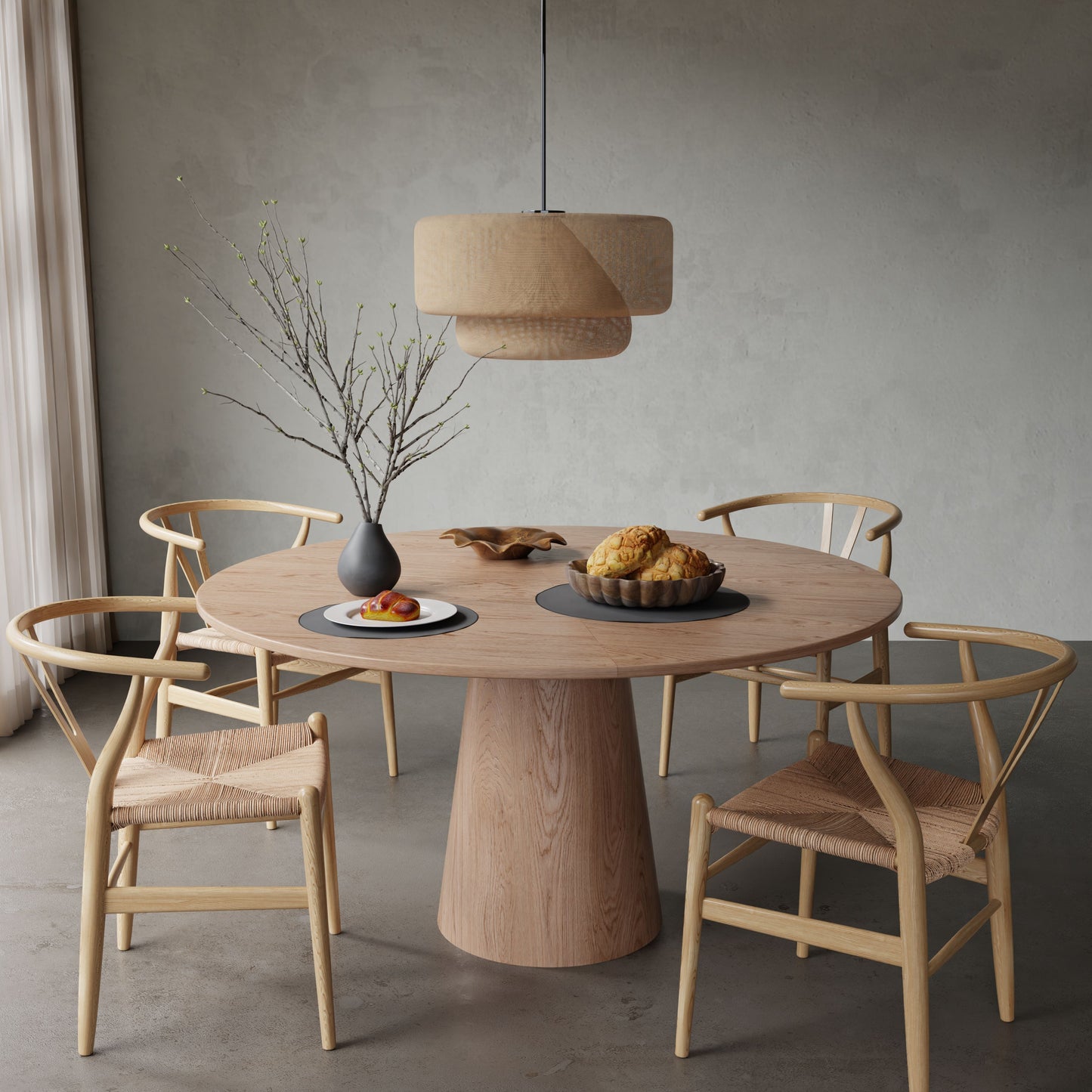 The Harlow + Elsa Dining Set, featuring a round wooden table and four woven chairs, sits in a minimal room beneath a woven pendant lamp. On the table are a vase with bare branches, fruit, bread, and pastries.
