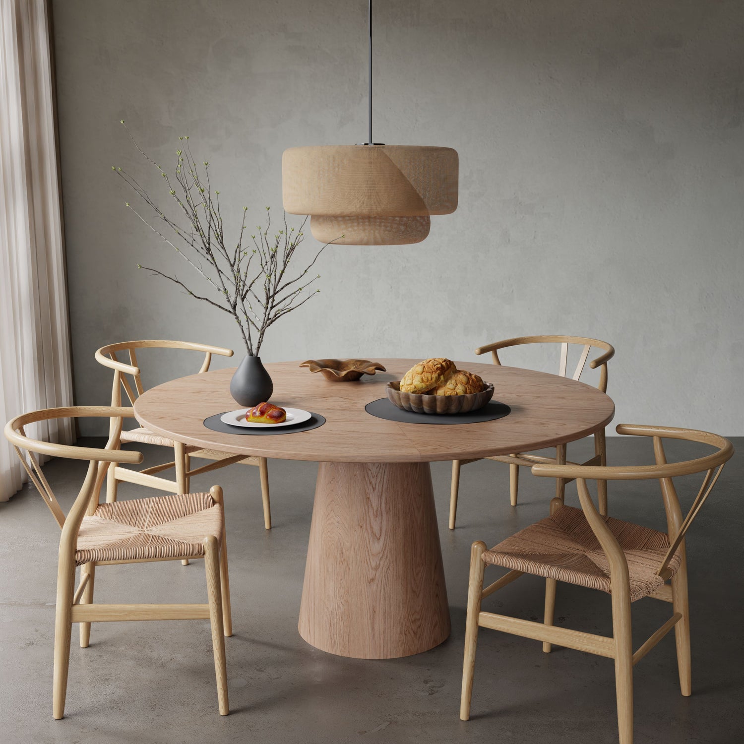 The Harlow + Elsa Dining Set, featuring a round wooden table and four woven chairs, sits in a minimal room beneath a woven pendant lamp. On the table are a vase with bare branches, fruit, bread, and pastries.