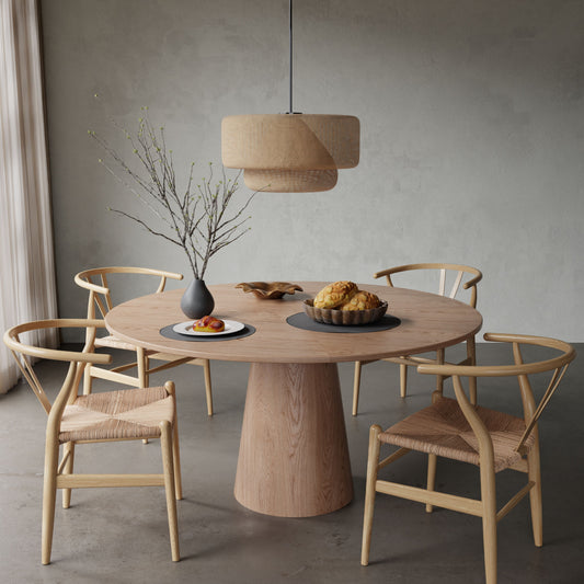 The Harlow + Elsa Dining Set, featuring a round wooden table and four woven chairs, sits in a minimal room beneath a woven pendant lamp. On the table are a vase with bare branches, fruit, bread, and pastries.