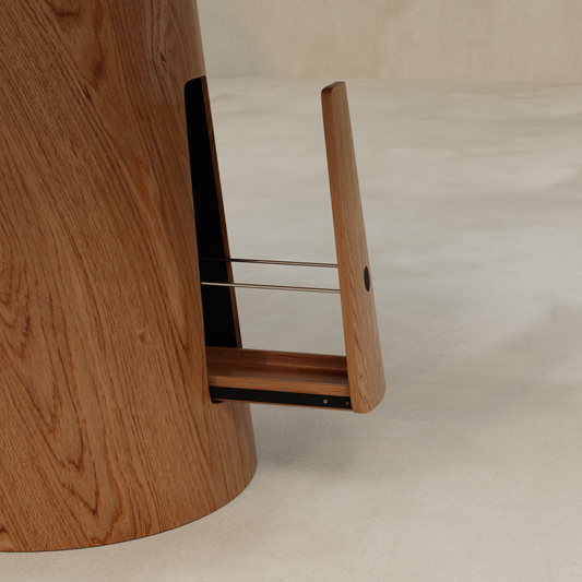 A close-up of the Nola Dining Table shows its cylindrical wooden frame with a hidden pull-out drawer—partially open to reveal metal rails and a smooth, light wood finish—standing on a beige floor.