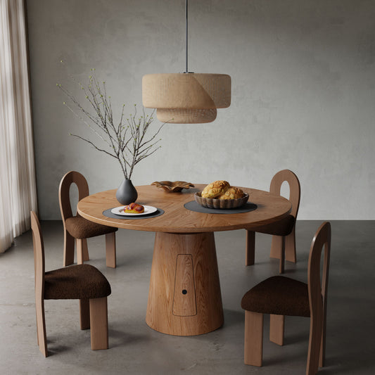The Nola + Aveline Dining Set features a round wooden table and four modern chairs arranged under a woven pendant light, accented with a vase of branches, and plates of pastries, fruit, and bread in a softly lit minimalist dining room.