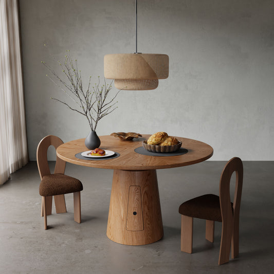 The Nola + 2 Aveline Dining Set features a round wooden table between two modern chairs, styled with a vase, fruit, and pastries. A beige pendant lamp hangs above while soft light filters through sheer curtains.