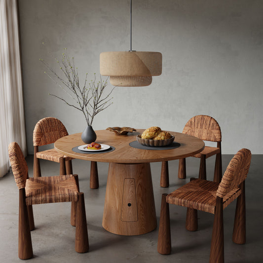 The Nola + Bardo Dining Set features a round wooden table and four woven chairs in a minimalist room. A gray vase with bare branches, pastry plate, and bread top the table, while a woven pendant lamp filters light through sheer curtains above.