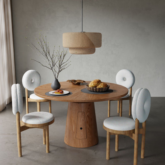 The Nola + Donut Dining Set features a round wooden table, four white cushioned chairs, a vase with branches, pastries on a tray, and a pendant lamp above, all set against a neutral gray wall for a modern dining space.