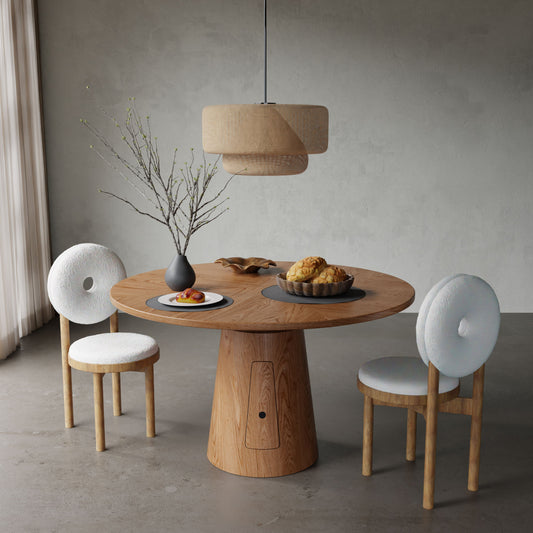 The Nola + 2 Donut Dining Set features a modern round wood table with two white cushioned chairs, styled with a black vase, fruit plate, and pastries. A beige pendant lamp hangs above in a minimalist gray-walled, gray-floored room.