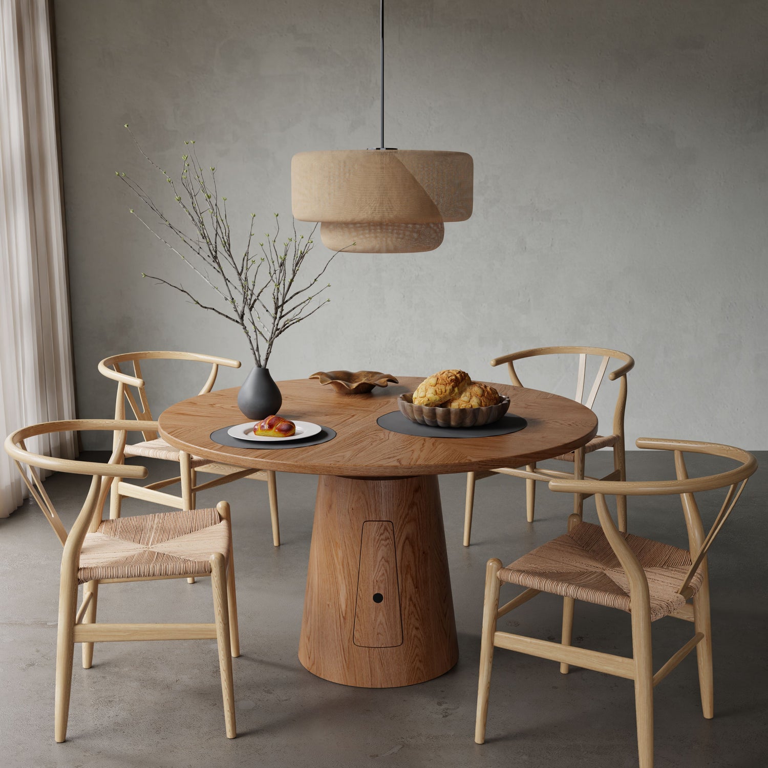 The Nola + Elsa Dining Set features a round wooden table with four woven chairs, complemented by a black vase with branches, pastries on a plate, and a pendant light. The minimalistic scene has neutral tones and gentle natural light.