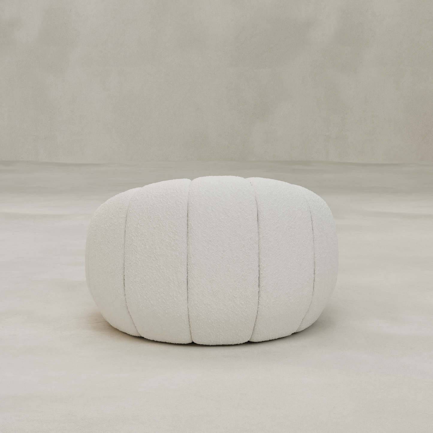 A round, white, textured pouf with vertical seams sits on a smooth, light-colored floor against a neutral backdrop, perfect for pairing with the Penelope Chair to create a cozy reading nook.