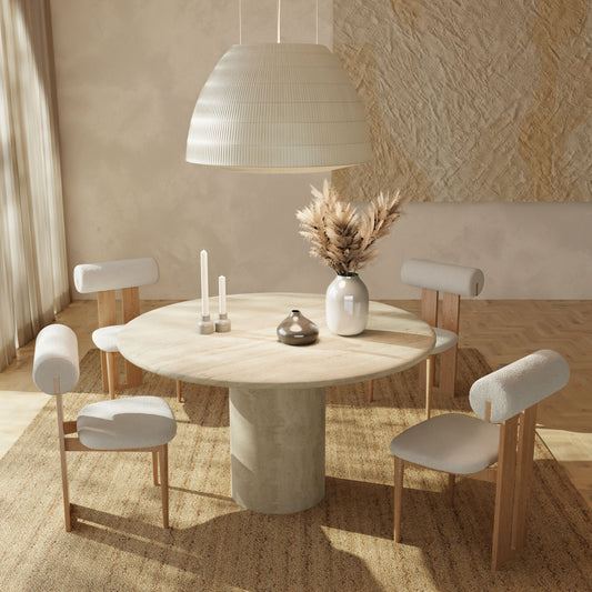 A minimalist dining room featuring the Renè + Templeton Dining Set—a round travertine pedestal table with four bouclé chairs—plus a large pendant lamp, candles, and a ceramic vase with pampas grass. Sunlight from the left highlights the neutral decor.
