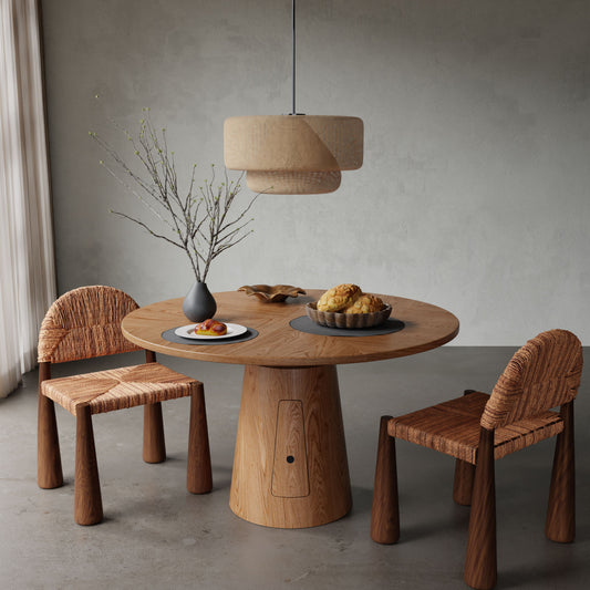 The Nola + 2 Bardo Dining Set features a round wood table with two woven chairs, complemented by a gray vase with branches, plate of pastries, and pendant lamp in a minimalist room with gray walls and floor.
