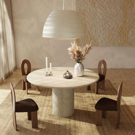 The Renè + Aveline Dining Set, featuring a round stone table and four wooden chairs on a woven rug, is styled with two white candles, a ceramic bowl, and a vase of dried grasses. Overhead, a large white pendant light completes the look.