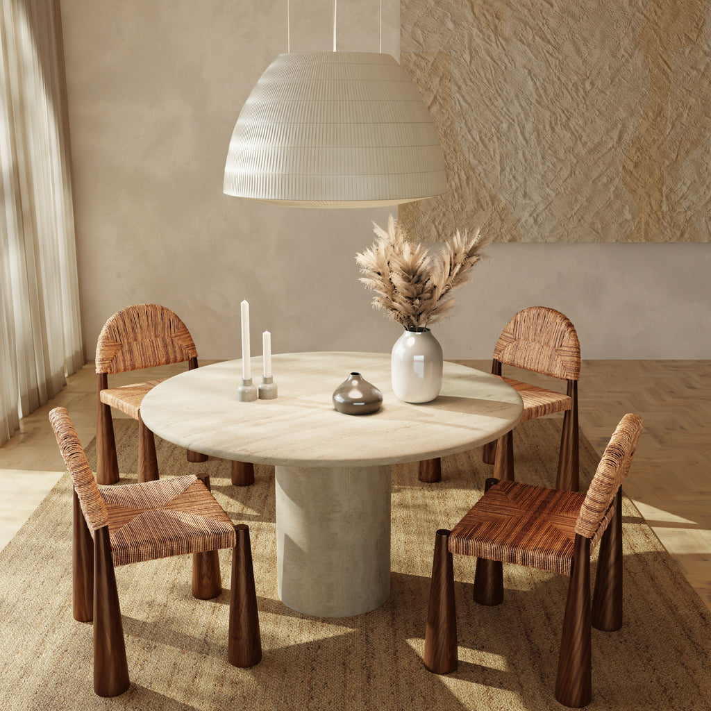 The Renè + Bardo Dining Set features a round table with a light stone base, four woven wooden chairs, two white candles, a ceramic dish, dried grasses in a vase, and is illuminated by a large white pendant lamp above.