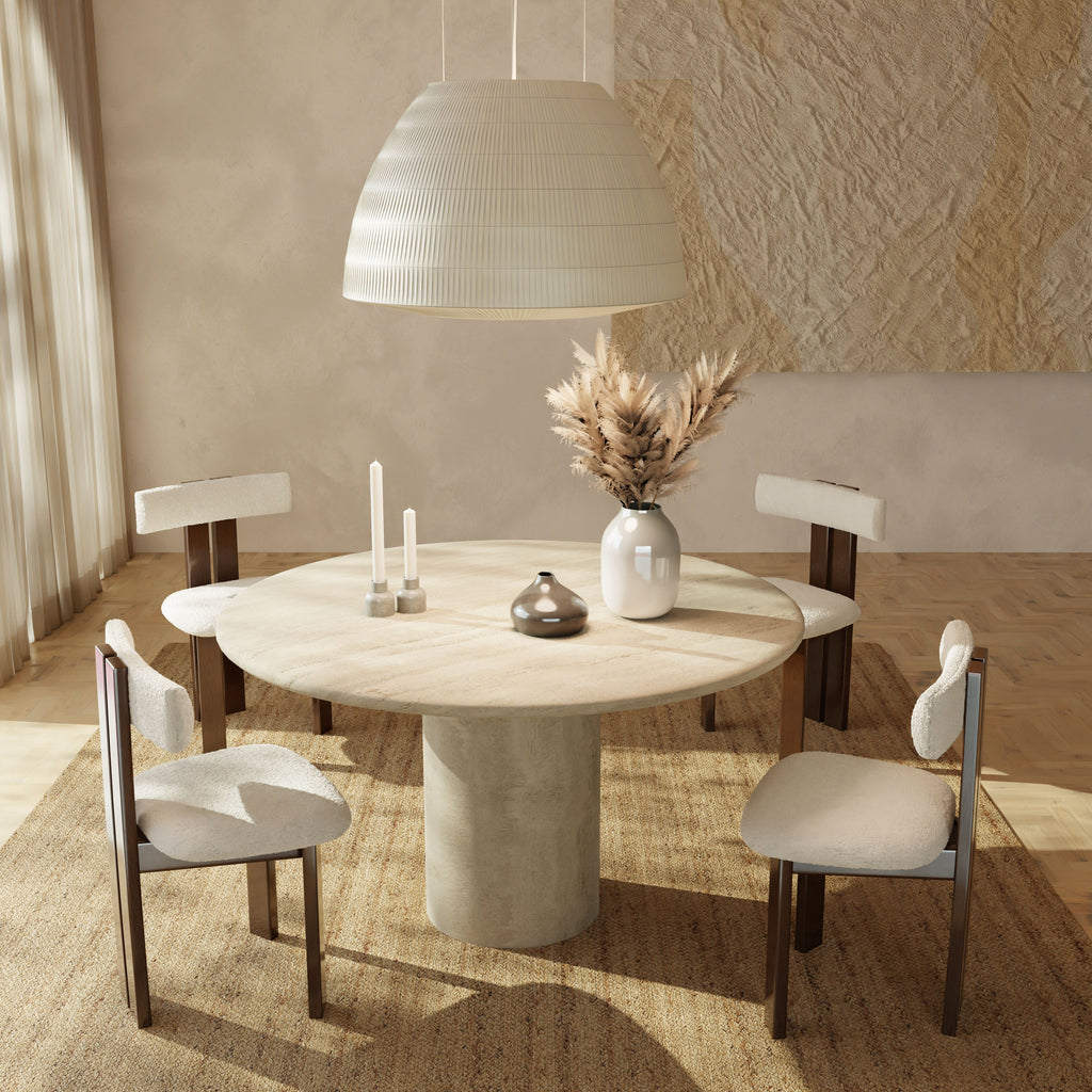 The Renè + Radley Dining Set features a round beige table and four modern chairs, styled with a white vase of pampas grass, candles, and decor on a textured rug beneath a large pendant in a bright minimalist room.