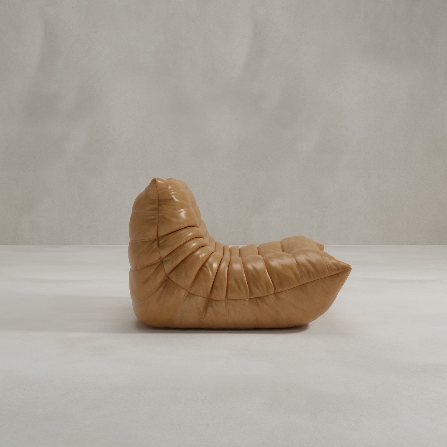 The Rolo Lounge Chair, featuring 70s-inspired design with tan leather, a low arched back, and deep pleated upholstery, sits on a smooth light gray floor against a plain textured gray wall.