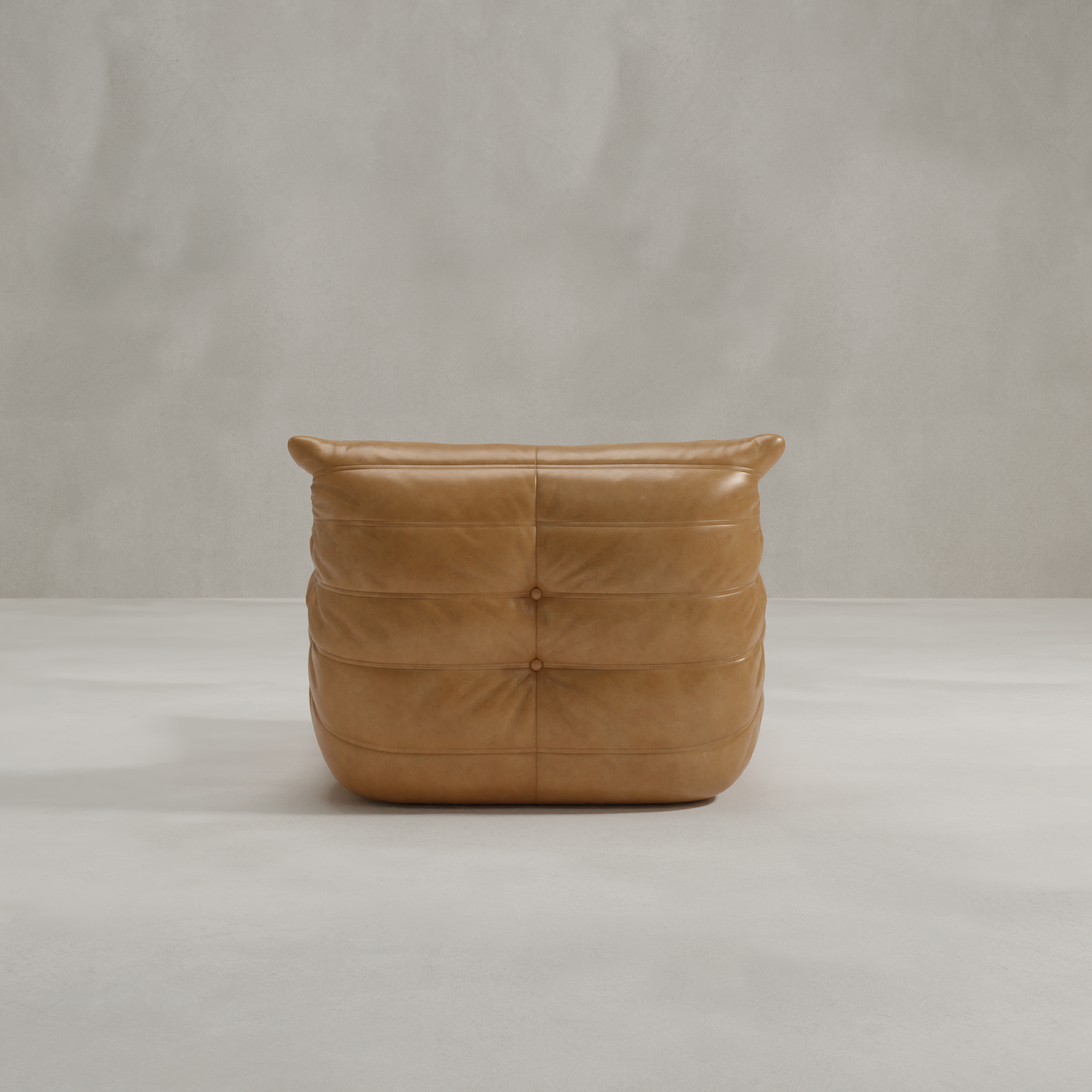 The Rolo Lounge Chair, featuring tan leather and tufted cushions, sits on a smooth light gray floor against a textured gray wall. Its low, wide silhouette and relaxed vibe evoke classic mid-century style.