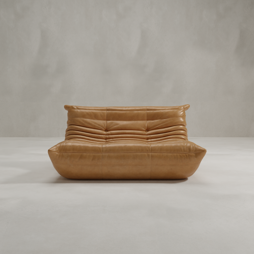 The Rolo Lounge Loveseat, featuring tan leather and mid-century style with plush ruched cushions, sits on a minimalist concrete floor against a plain light gray wall.