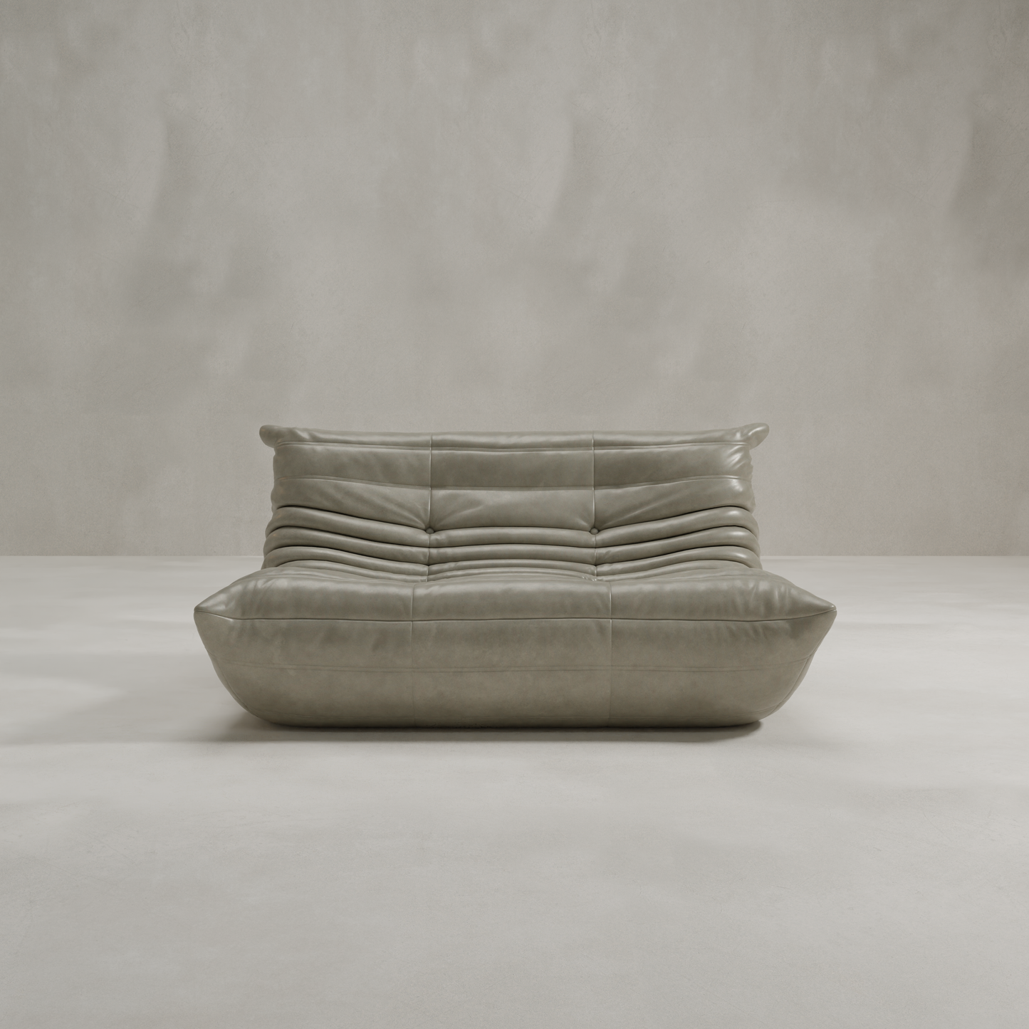 The Rolo Lounge Loveseat, a modern low-profile piece with tufted gray leather and plush cushioning inspired by mid-century design, sits in a minimalist room featuring a light gray textured wall and floor.