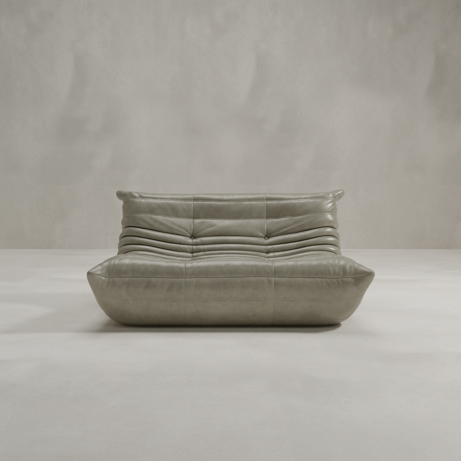 The Rolo Lounge Loveseat, a modern low-profile piece with tufted gray leather and plush cushioning inspired by mid-century design, sits in a minimalist room featuring a light gray textured wall and floor.