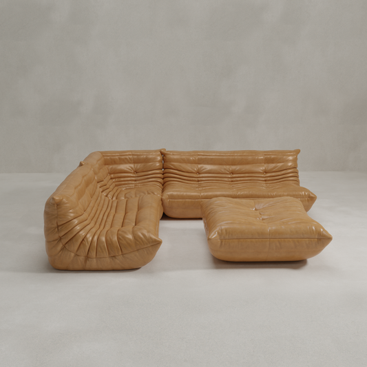 The Rolo Lounge Four Piece Sofa in tan leather, featuring tufted cushions and a low profile, sits on a light minimalist floor by a plain grey wall. A matching ottoman is placed at the center.