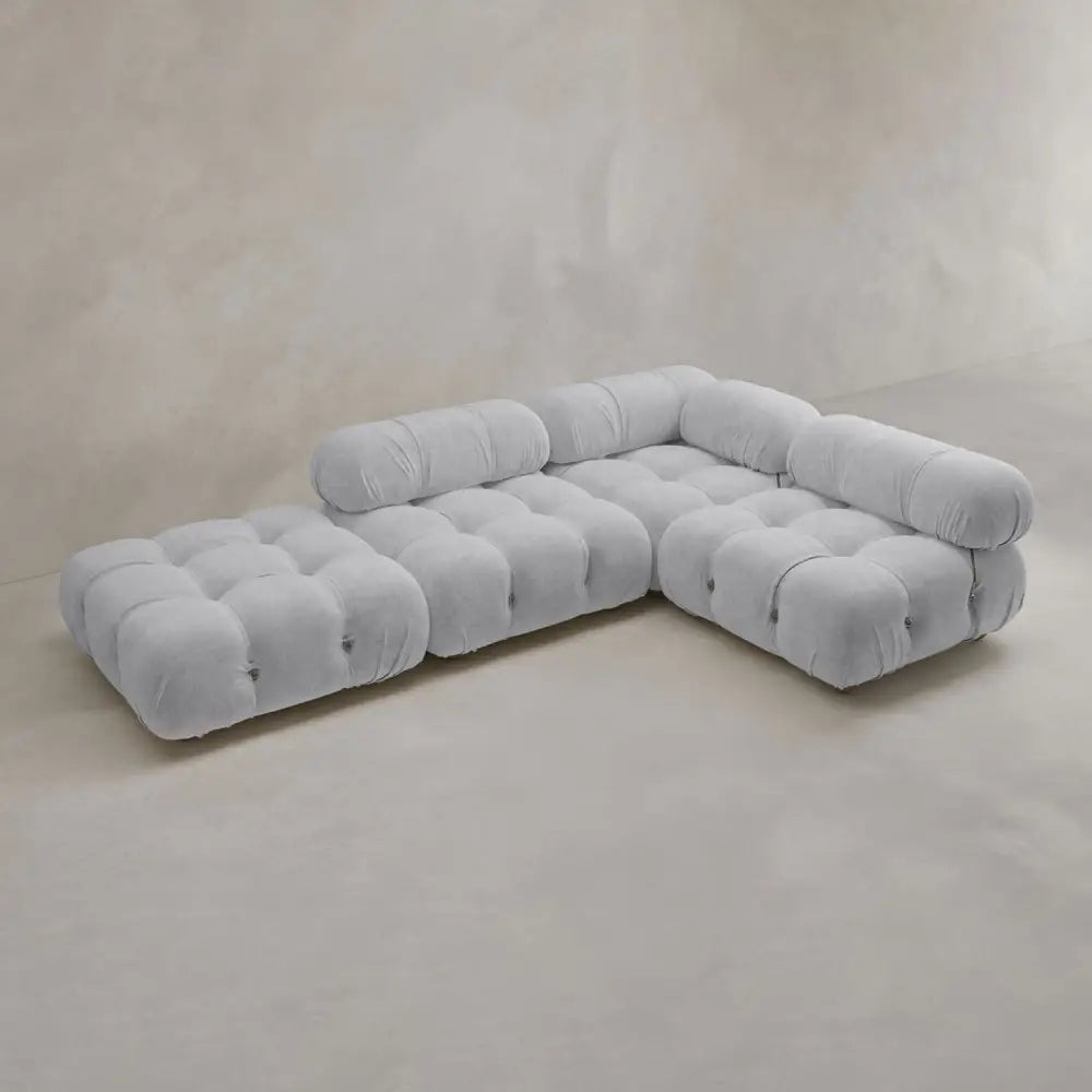 The Belmont Sofa Combination C is a modern, light gray tufted sectional with classic appeal, rounded armrests, and marshmallow-like curves, set on a smooth neutral-toned floor against a plain beige wall.