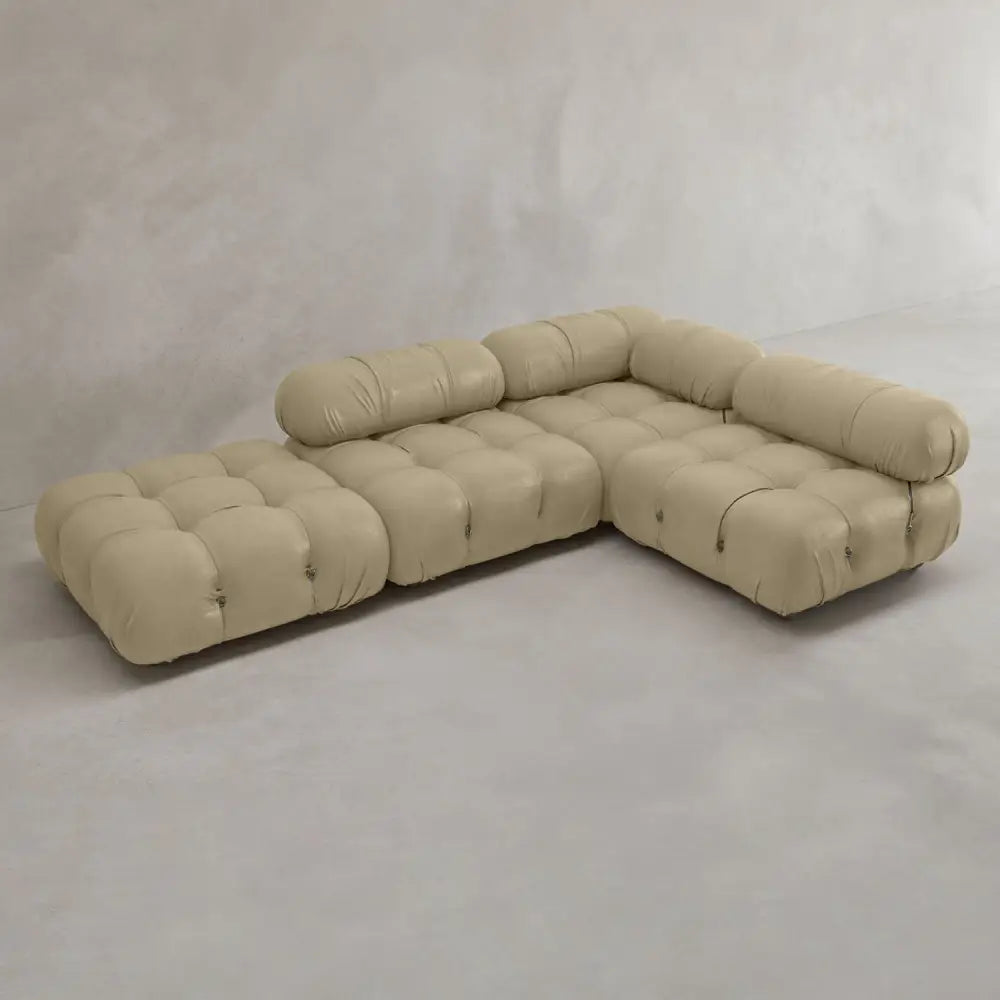 The Belmont Sofa Combination C is a modern beige sectional with tufted cushions and soft, curved lines in an L-shape, set in a minimalist room with light gray walls and flooring.