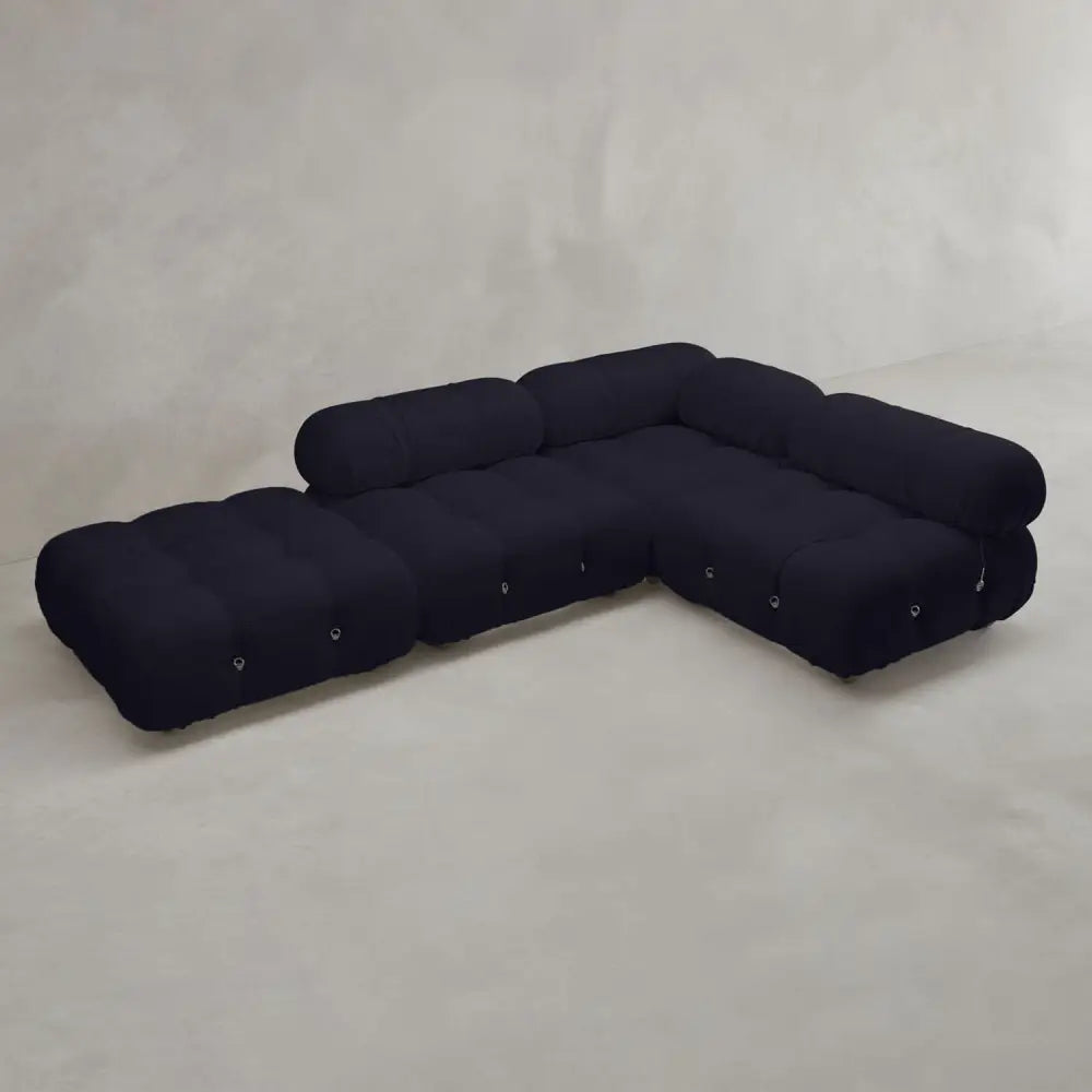 The Belmont Sofa Combination C is a modern black modular sectional with marshmallow-like curves, rounded tufted cushions, and low backrests. It sits on a light gray floor against a plain, textured wall.