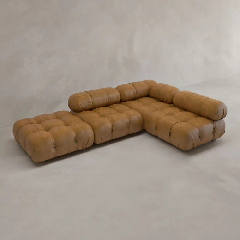The Belmont Sofa Combination C is a modular brown leather sectional with marshmallow curves, tufted design, thick cushions, and a low L-shape placed on a light gray floor against a plain light wall.