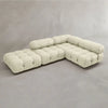 The Belmont Sofa Combination C is a modern, modular cream-colored sectional with tufted cushions and cylindrical backrests, arranged in an L-shape against a light grey wall and floor.