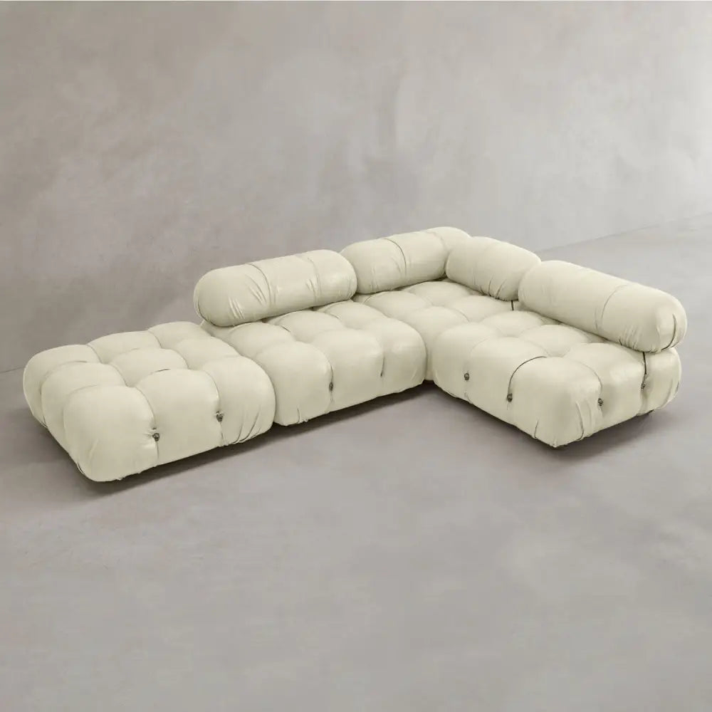 The Belmont Sofa Combination C is a modern, modular cream-colored sectional with tufted cushions and cylindrical backrests, arranged in an L-shape against a light grey wall and floor.