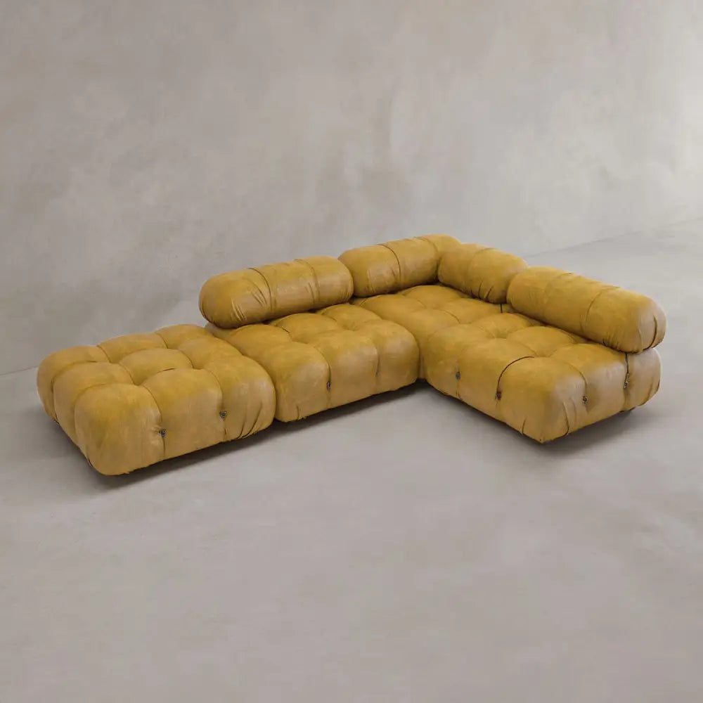 The Belmont Sofa Combination C is a modular, mustard yellow sectional with classic tufting and marshmallow-like curves, featuring low backrests and seat cushions in an L-shape on a light gray floor against a textured wall.