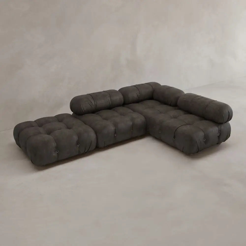 The Belmont Sofa Combination C, a low-profile dark gray modular sectional with tufted cushions and soft curves, is arranged on a light concrete floor against a plain neutral wall.