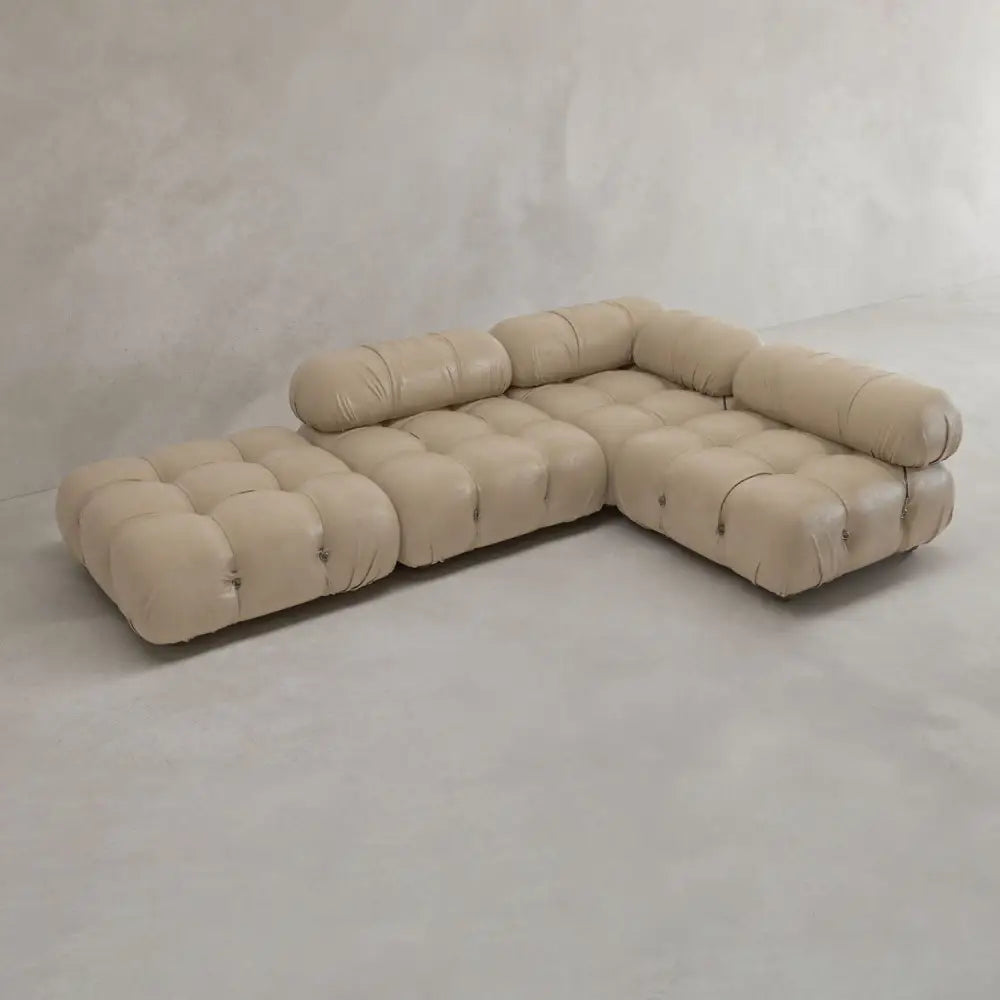 The Belmont Sofa Combination C, a modern light beige modular sectional with marshmallow-like curves and tufted cushions, sits on a smooth light gray floor against a textured off-white wall—an iconic classic of modular design.