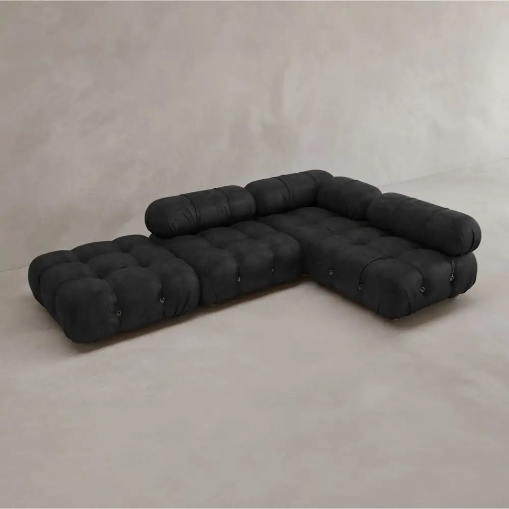 The Belmont Sofa Combination C is a modern, modular black sectional with tufted cushions, marshmallow-like curves, and cylindrical bolsters, arranged in an L-shape against a plain light-colored wall and floor.