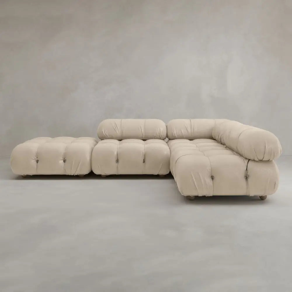 The Belmont Sofa Combination C, a modern, modular beige sectional with tufted cushions and rounded edges, features marshmallow-like curves and sits on a light gray floor against a neutral gray wall.