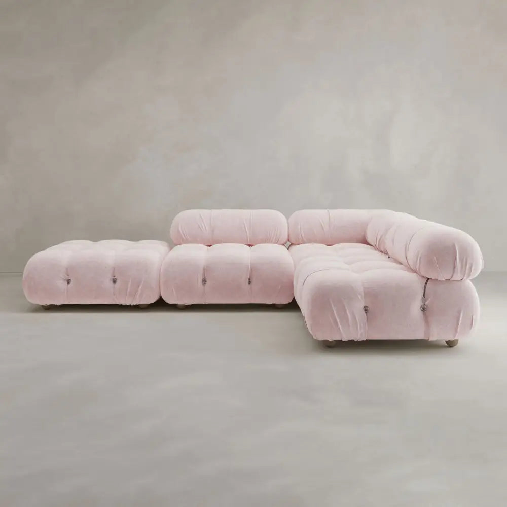 The Belmont Sofa Combination C, a modern modular sectional in light pink with marshmallow-like curves and tufted cushions, features a chaise lounge and matching ottoman, positioned against a plain textured grey background.