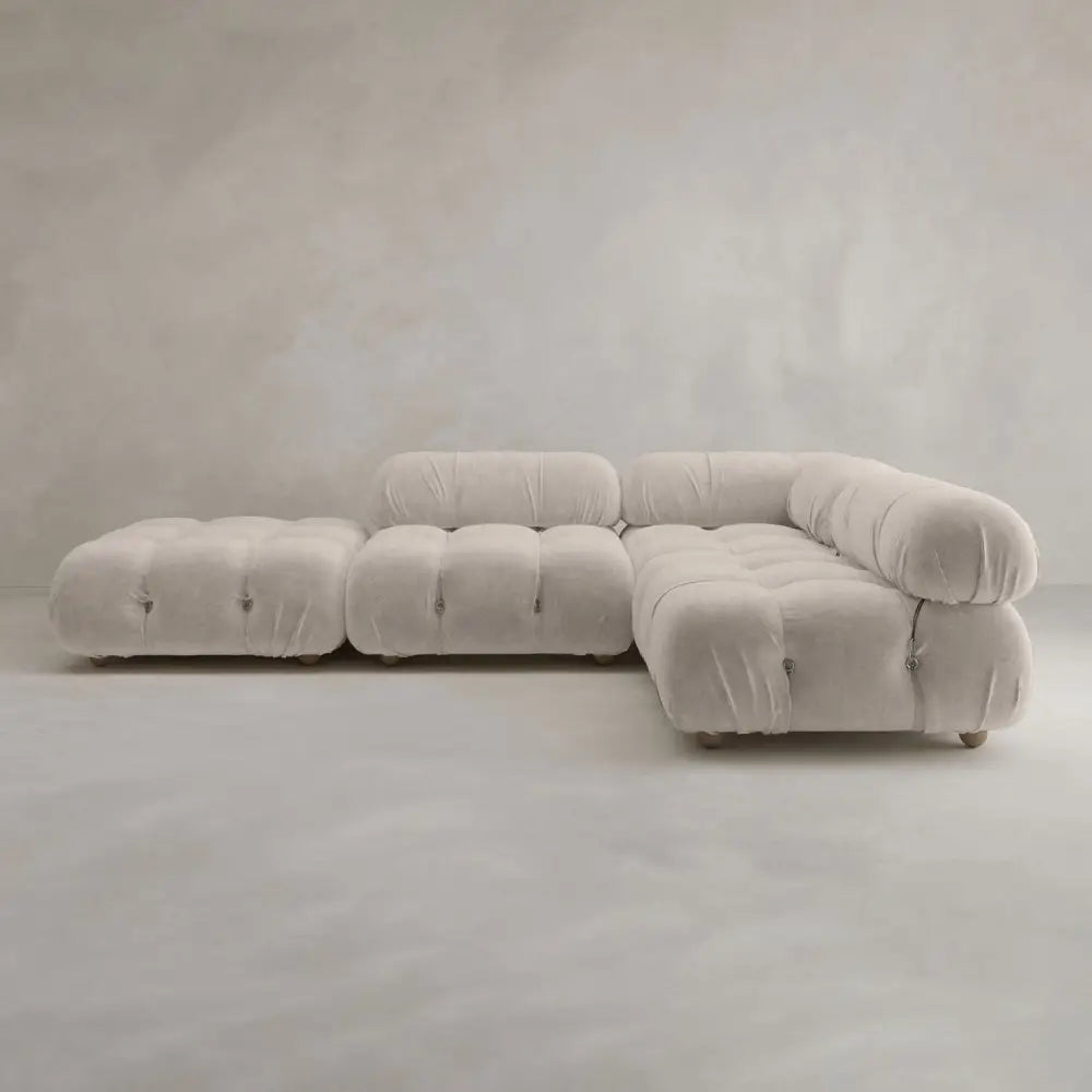 The Belmont Sofa Combination C is a modern, light beige sectional with tufted, marshmallow-like curves and an ottoman, set against a neutral wall and floor.