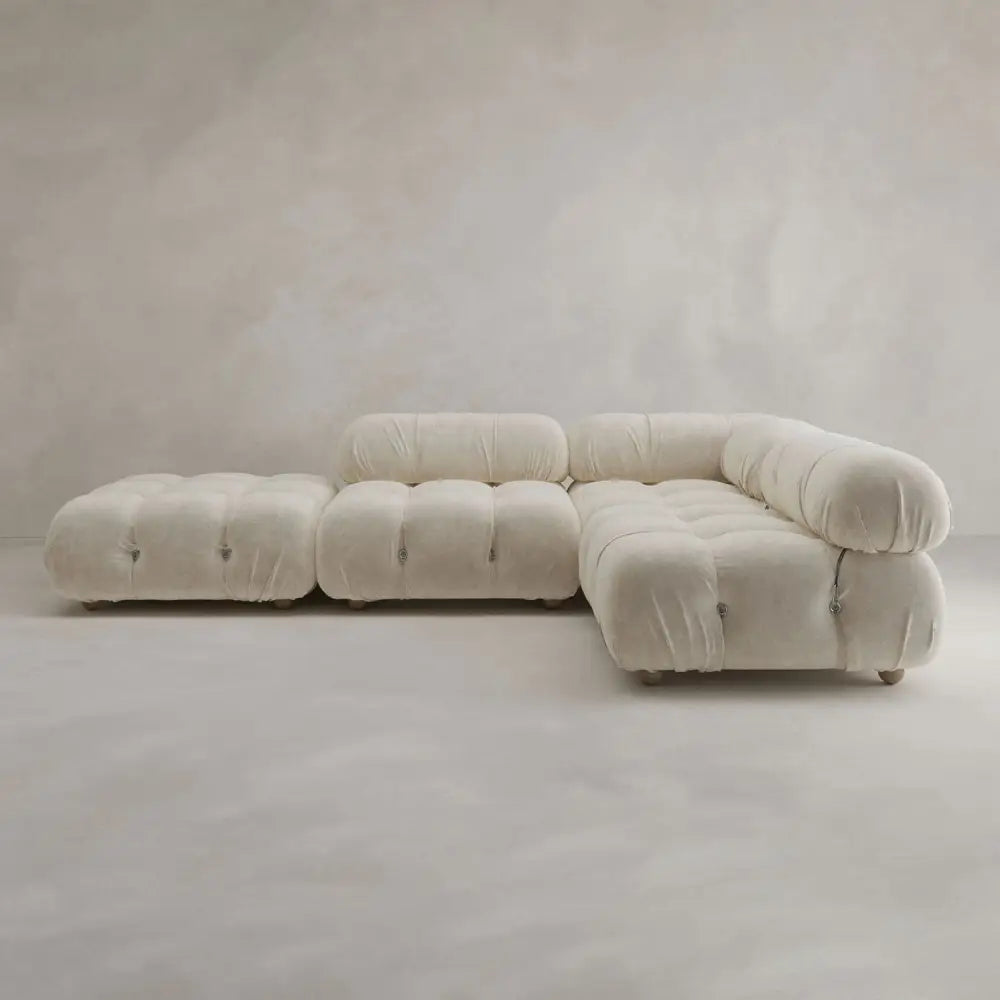 The Belmont Sofa Combination C is a modern modular sectional with marshmallow-like curves, tufted rounded cushions, and a matching ottoman, all upholstered in light beige fabric and set against a plain, light background.