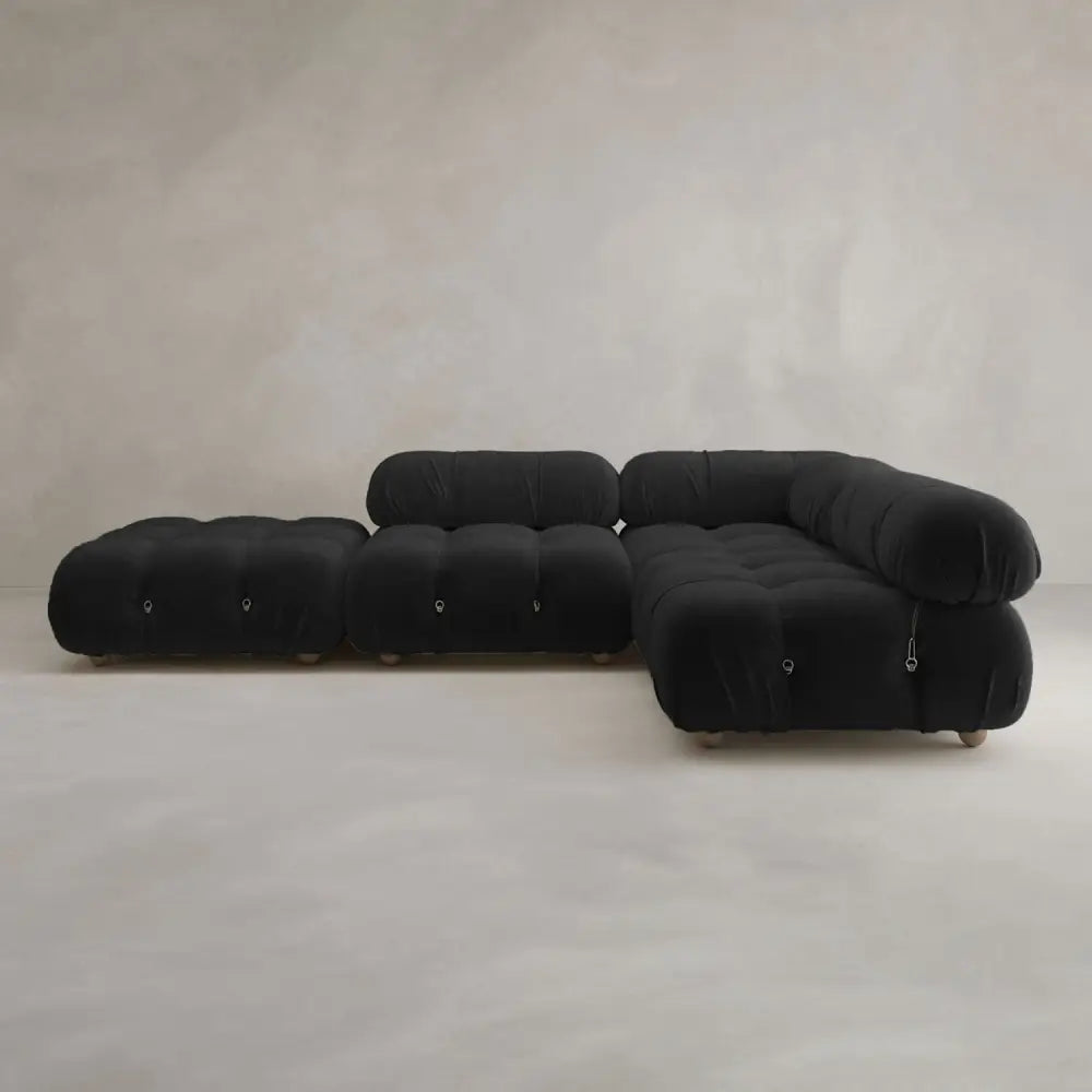 The Belmont Sofa Combination C, a modern black sectional with curved, tufted cushions, sits next to a matching ottoman against a textured neutral wall and light floor.