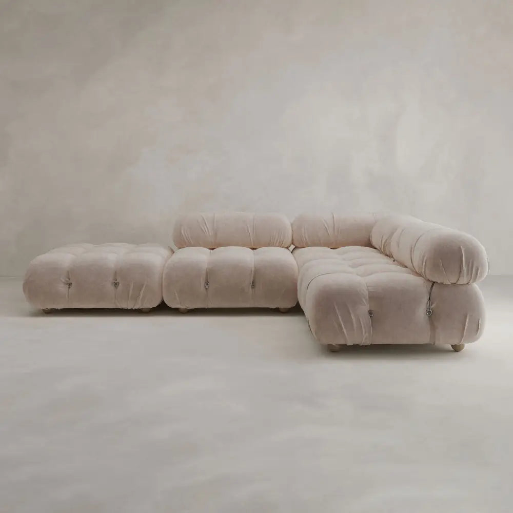 The Belmont Sofa Combination C, a modern modular beige sectional with plush tufted cushions and rounded curves, sits in a minimalist room featuring light, textured walls and a matching floor.