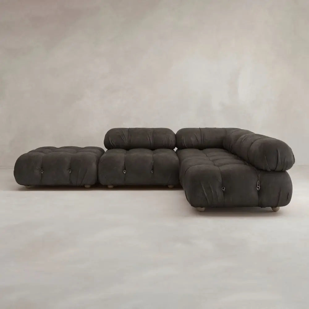 The Belmont Sofa Combination C is a modular, dark gray tufted sectional with marshmallow-like curves and an ottoman, set against a plain light-colored wall and floor.