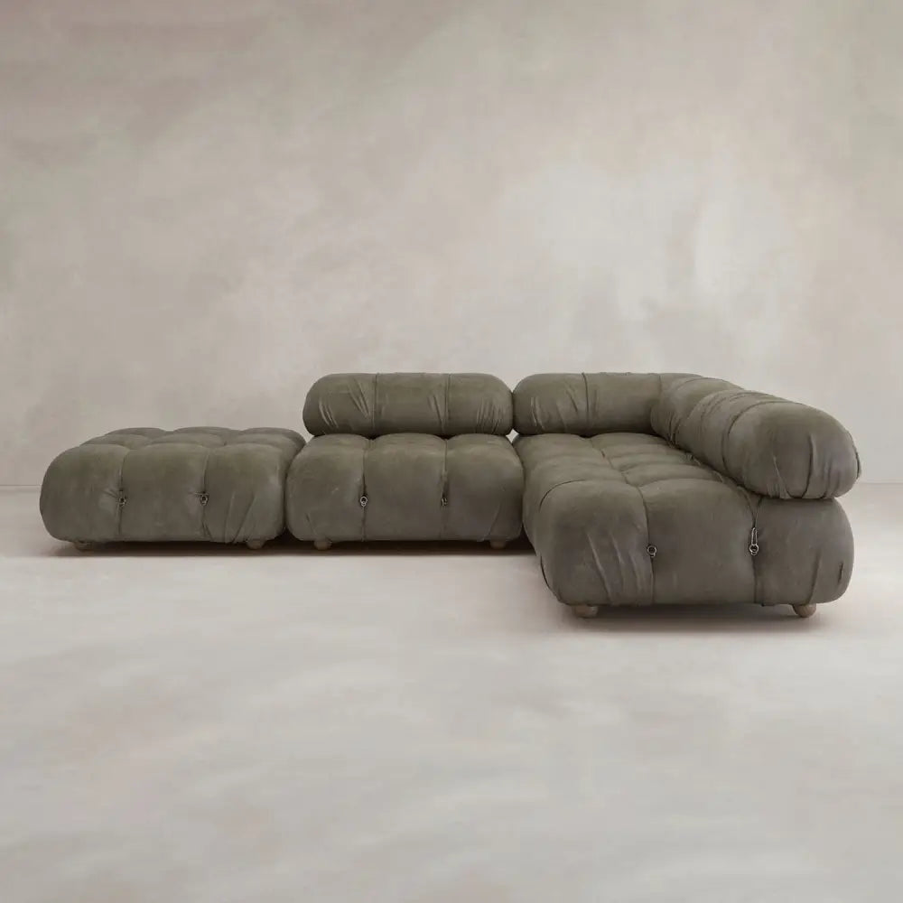 The Belmont Sofa Combination C is a modern olive-green sectional with marshmallow-like curves, tufted cushions, and rounded edges. Its modular design features a chaise lounge and matching ottoman, set against a neutral textured wall and light floor.