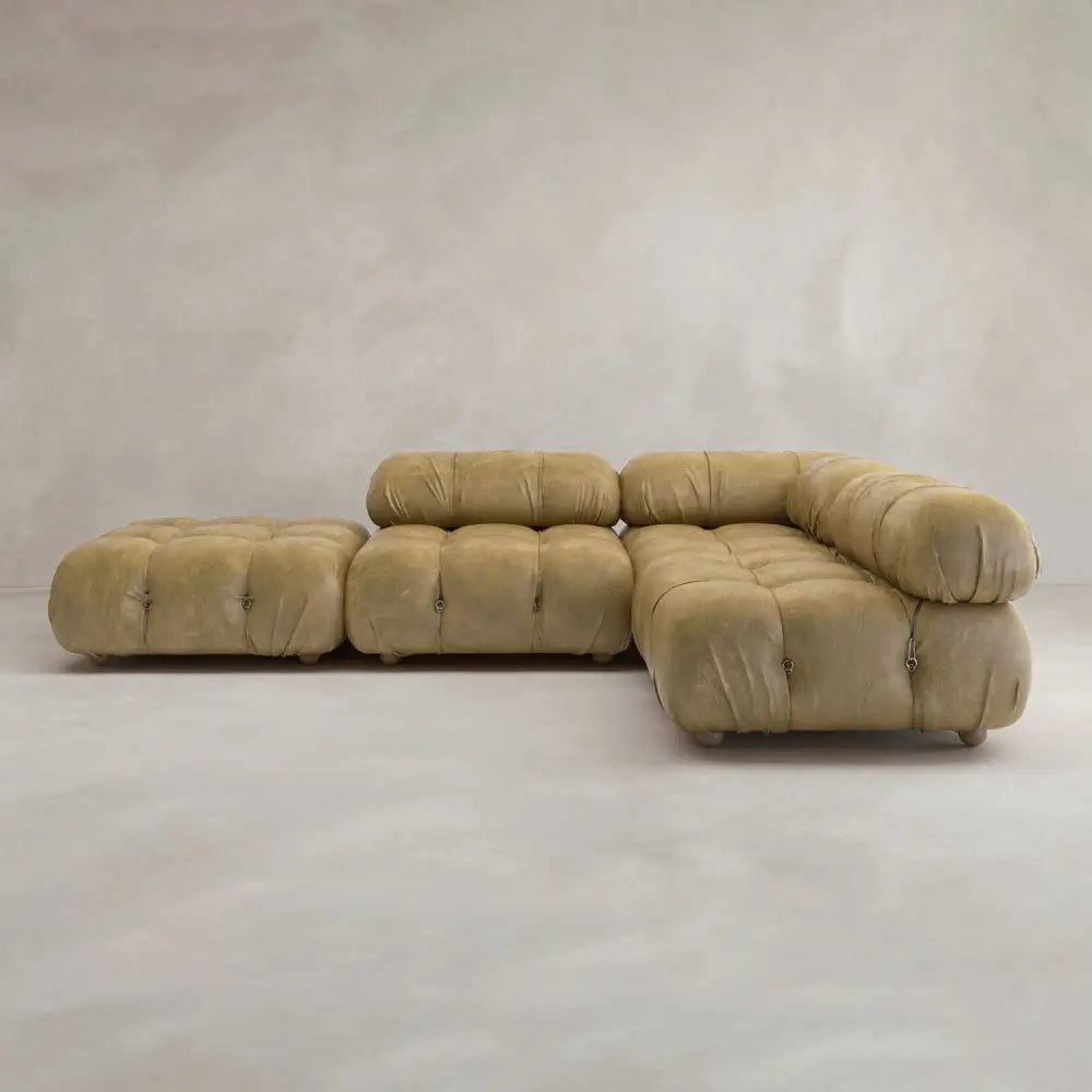 The Belmont Sofa Combination C is a modular, tufted olive-green sectional with marshmallow-like curves, an attached chaise lounge, and a matching ottoman, set against a plain light beige wall and floor.