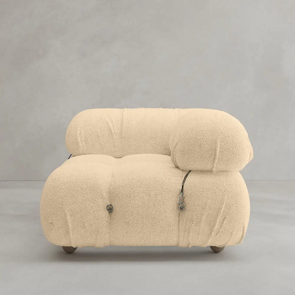 The Belmont Sofa Left Armrest features a modern modular design with soft, textured beige fabric, rounded edges, visible tufting, and exposed metal accents, shown against a plain gray background.