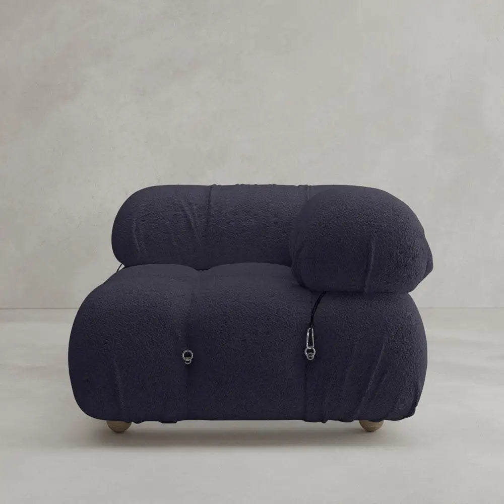 The Belmont Sofa Left Armrest features a modern, deep navy look with plush, rounded design and soft, textured fabric. Inspired by classic styles, it has metal ring and zipper accents on the sides against a plain light gray background.