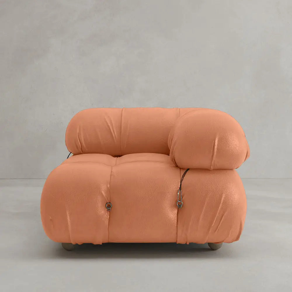 The Belmont Sofa Left Armrest, featuring plush marshmallow-like curves and soft orange upholstery with visible button tufts and metal fasteners, sits against a plain light gray wall and floor background.