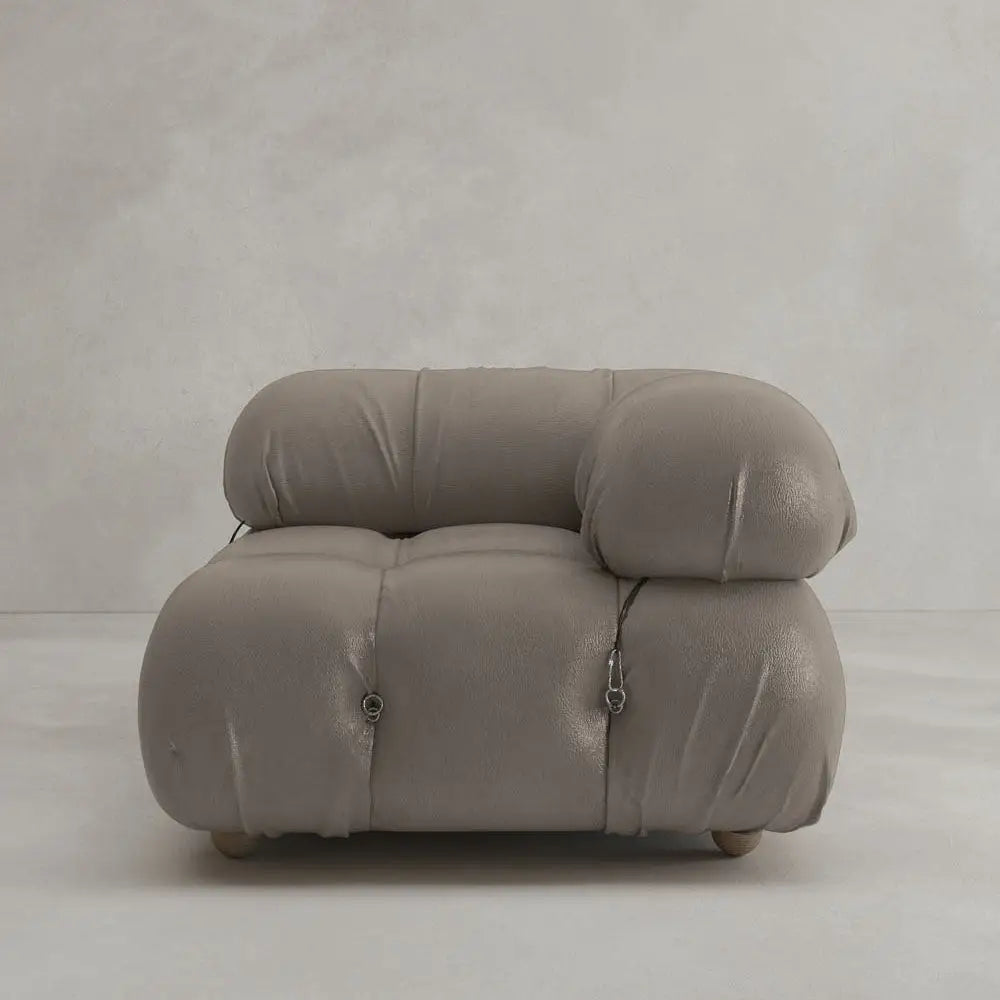 The Belmont Sofa Left Armrest in plush taupe, featuring a rounded, tufted design and visible seams, offers an iconic classic look against a plain light gray wall and floor.