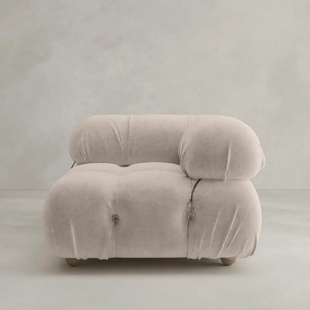 The Belmont Sofa Left Armrest features plush, light beige upholstery with marshmallow-like curves, subtle button tufting, and small wooden legs, perfect against a neutral textured wall and floor.